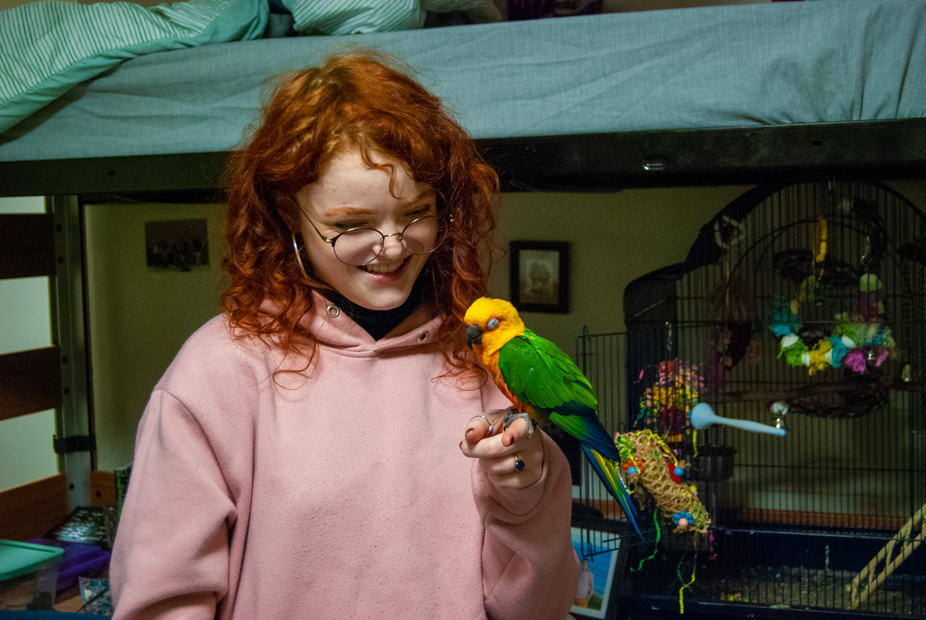 India holds Ollie on her finger and smiles at him. Ollie's cage and toys are in the background.