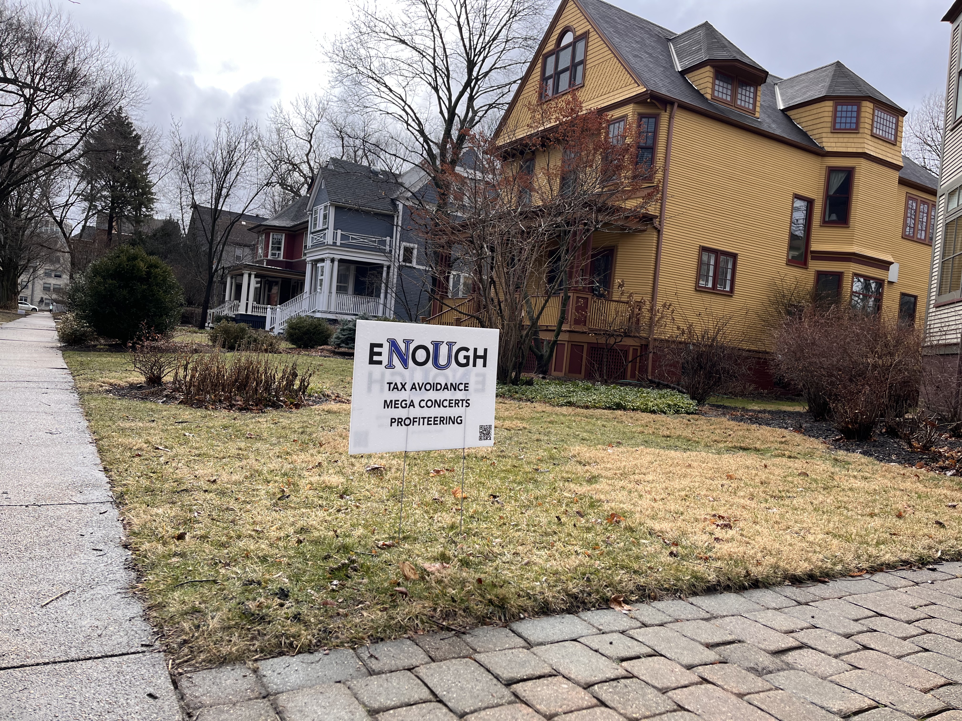 A yard sign staked in front of a yellow house that says 'Enough tax avoidance, mega concerts, profiteering.'