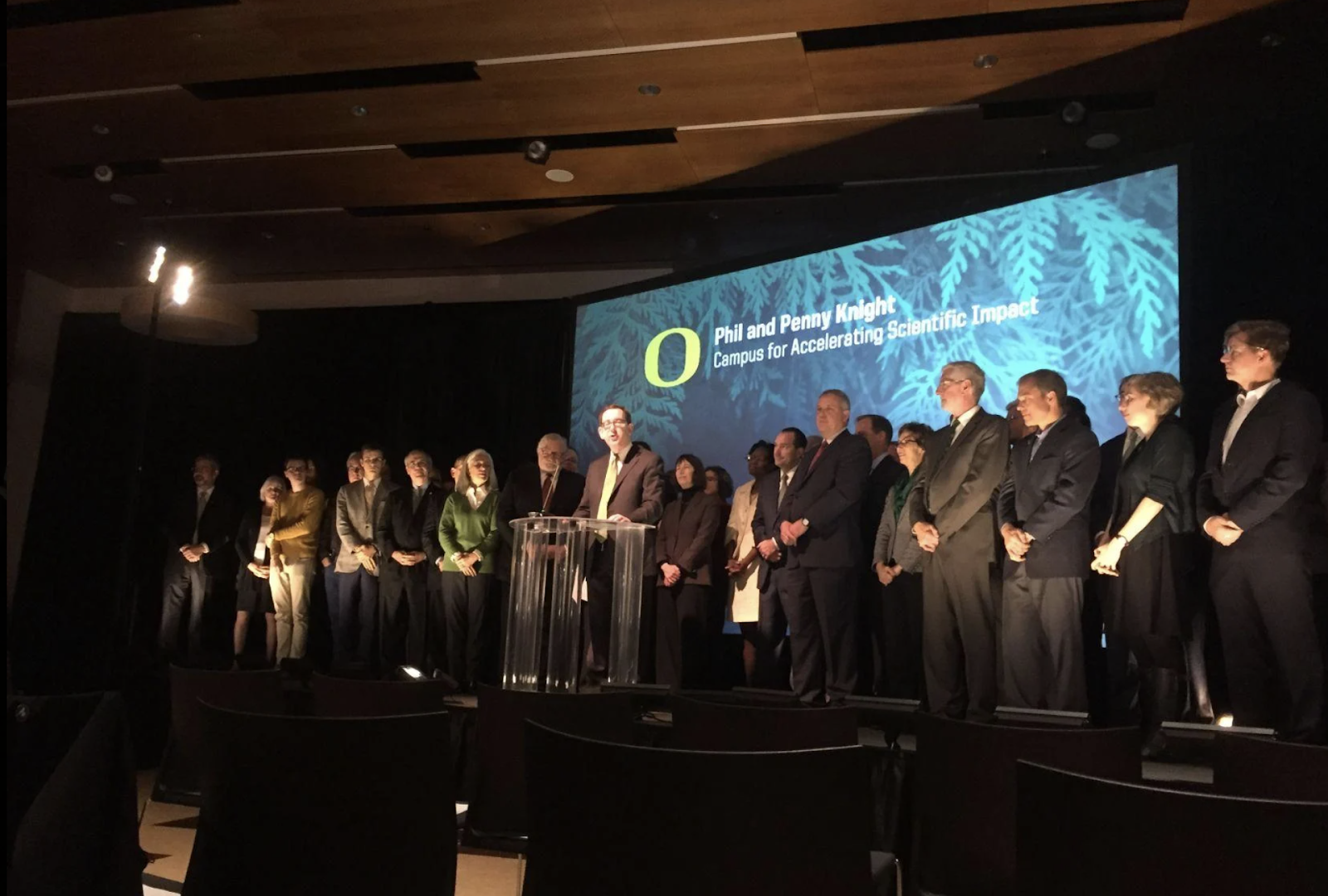 Schill stands before a podium on stage at the University of Oregon, surrounded by faculty members and other stakeholders.